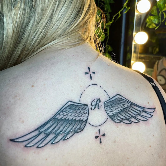 Medium Black and Grey Angel Wings with R and Dots Tattoo on Women Shoulder by @chxrlieseye.ttt