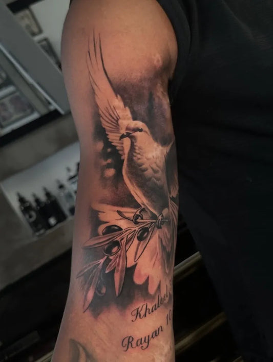Medium Black and Grey Dove with Olive Branch and Name Tattoo on Men Arm by @southgatetattoo