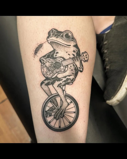 Medium Black and Grey Frog on Unicycle with Ukulele Tattoo on Women Leg by @liv.laeta.tattoo