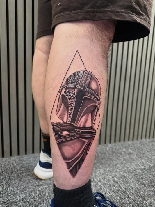 Medium Black and Grey Mandalorian Helmet Star Wars Tattoo on Men Leg by @kelseywalkertattoos