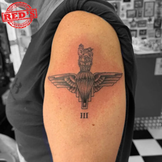Medium Black and Grey Parachute Wings with Crown and Roman Numerals Military Tattoo on Women Upper Arm by @redstattooparlour