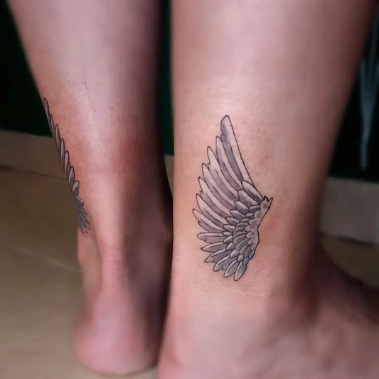 Small Black and Grey Angel Wings Tattoo on Women Ankle by @rona.m.tattoo