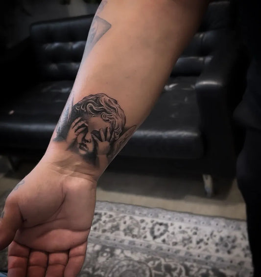 Small Black and Grey Cherub Tattoo on Women Forearm by @aroncowlestattoo