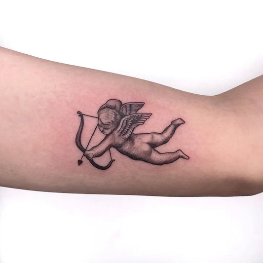 Small Black and Grey Cherub with Bow Tattoo on Women Arm by @kozie.tn