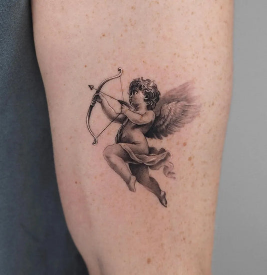 Small Black and Grey Cherub with Bow Tattoo on Women Arm by @oscarjesustattoo
