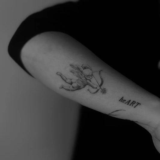 Small Black and Grey Cherub with Bow and Heart Tattoo on Women Arm by @defneink