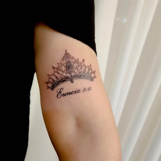 Small Black and Grey Crown with Script Tattoo on Women Upper Arm by @bada.tattoo