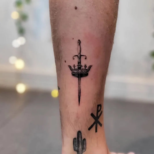 Small Black and Grey Crowned Sword and Chi Rho Tattoo on Men Leg by @tattoocrazy_cambridge