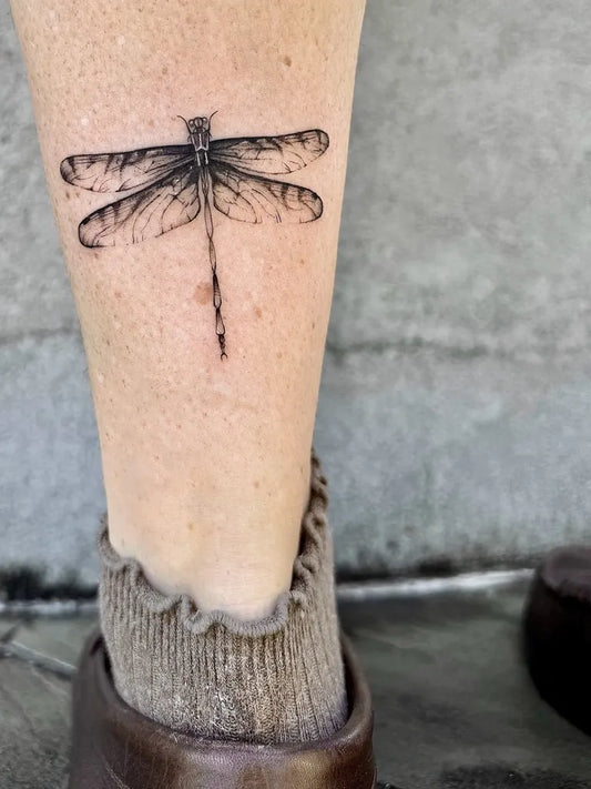Small Black and Grey Dragonfly on Women Leg Tattoo by @lyndsay.ink