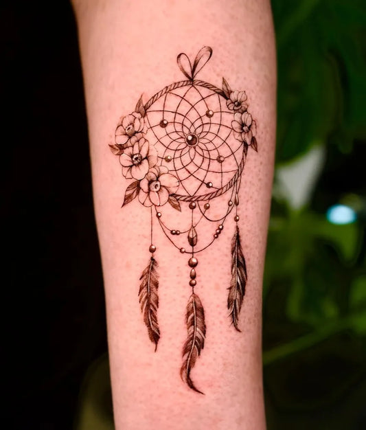 Small Black and Grey Dream Catcher with Flowers and Feathers Tattoo on Women Arm by @taeri_tattoo