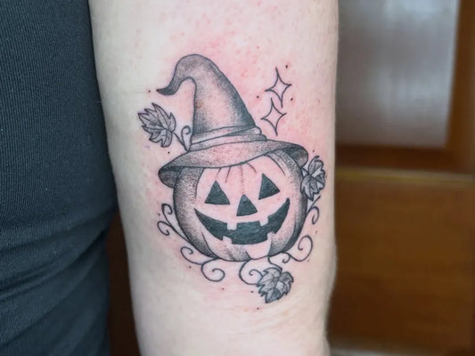 Small Black and Grey Jack o lantern with Witch Hat and Leaves Halloween Tattoo on Women Arm by @leah_wilt_tattoo