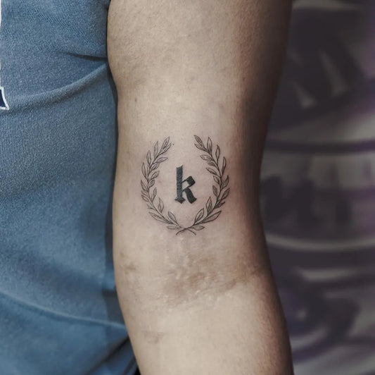 Small Black and Grey Laurel Wreath with Initial K Tattoo on Men Arm by @6.tattooist