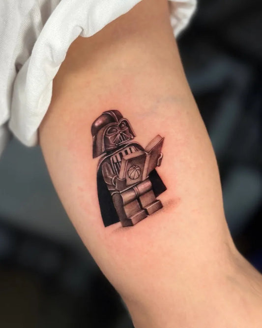 Small Black and Grey Lego Darth Vader Reading Tattoo on Women Upper Arm by @ruth.tatts