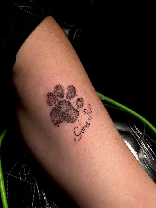 Small Black and Grey Paw Print with Name Tattoo on Women Upper Arm by @sicknesstattoos