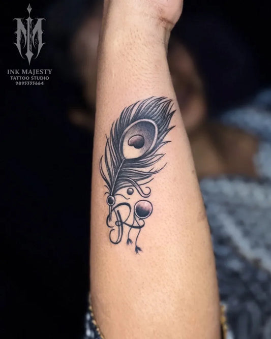 Small Black and Grey Peacock Feather with Initials Tattoo on Women Arm by @tattoo_ink_majesty_taliparamba