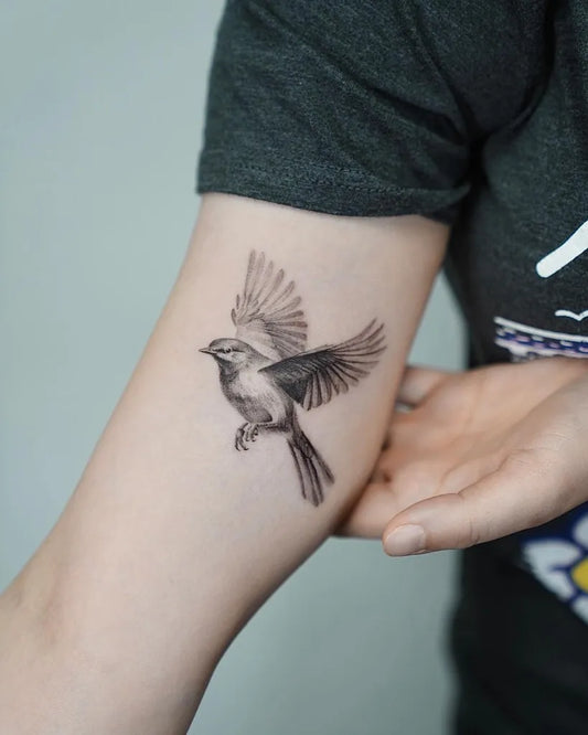 Small Black and Grey Sparrow Tattoo on Women Upper Arm by @baronarttattoo
