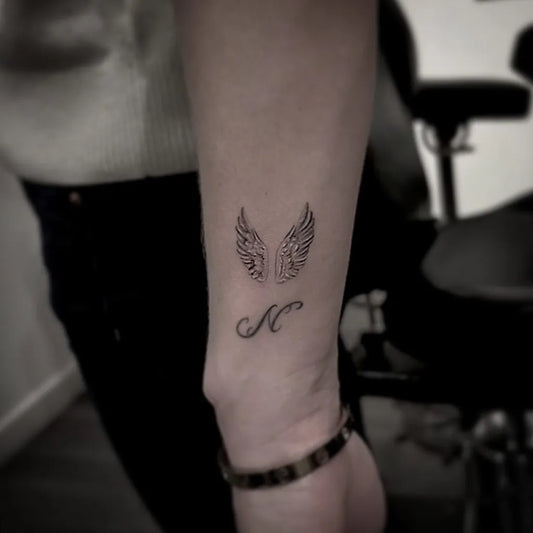 Small Blackwork Angel Wings and Initial Tattoo on Women Wrist by @aroncowlestattoo