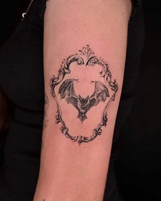 Small Blackwork Bat in Ornate Frame Tattoo on Women Upper Arm by @mercilyss.tattoos