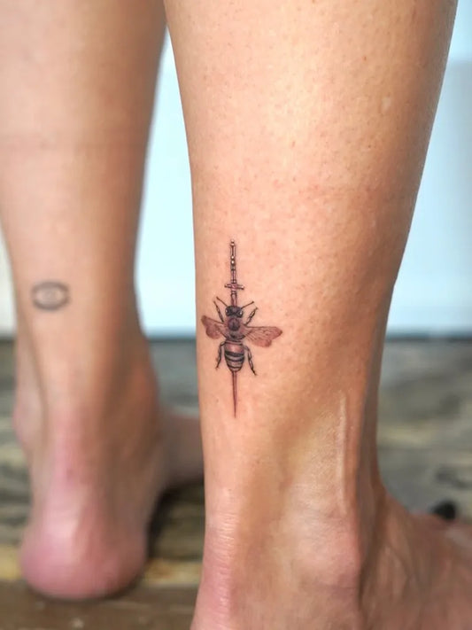 Small Blackwork Bee with Sword on Women Ankle Tattoo by @monochrometattoo
