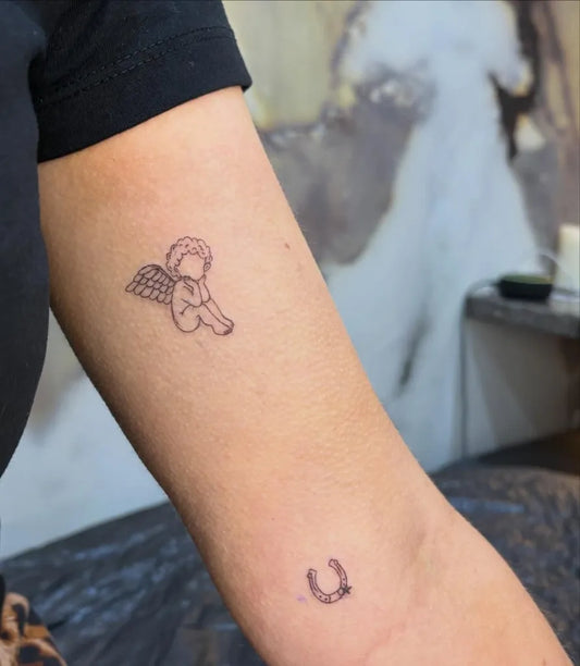 Small Fine Line Cherub and Horseshoe Tattoo on Women Arm by @blossomink__