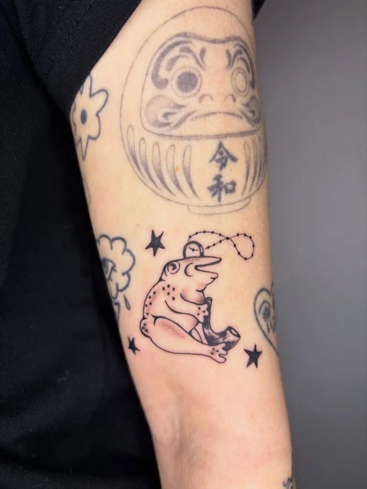 Small Fine Line Frog and Daruma Tattoo on Women Arm by @livluvstab