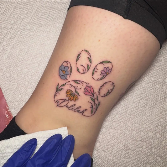 Small Fine Line Paw Print with Flowers and Name Tattoo on Women Leg by @tattysbynoodlez