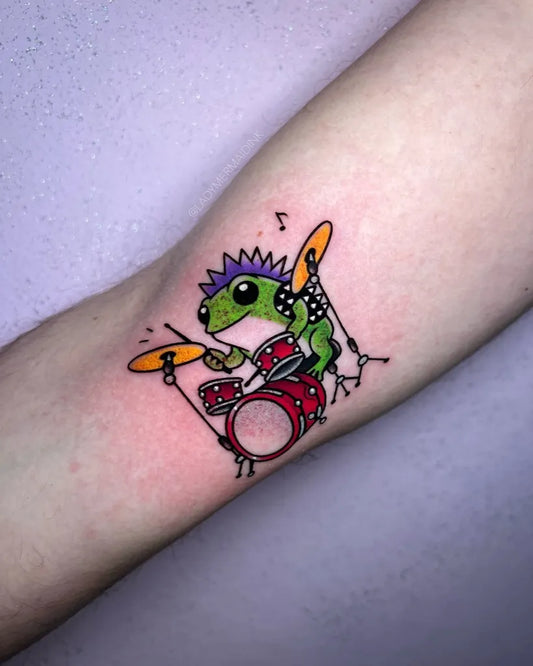 Small Neotraditional Chameleon Playing Drums Tattoo on Women Arm by @ladymermaidink