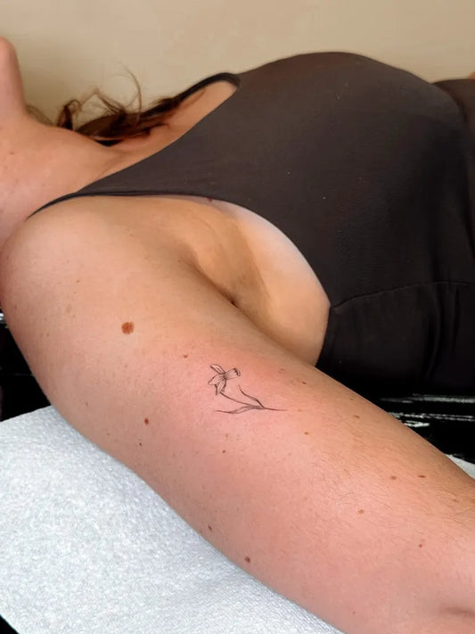 Tiny Fine Line Daffodil Tattoo on Women Upper Arm by @sorcha.elspeth.tattoo