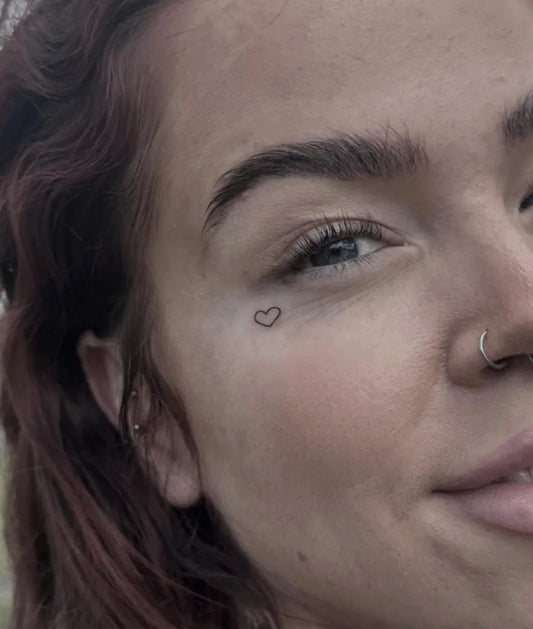 Tiny Minimalist Heart on Women Face Tattoo by @tomharrilltattoos