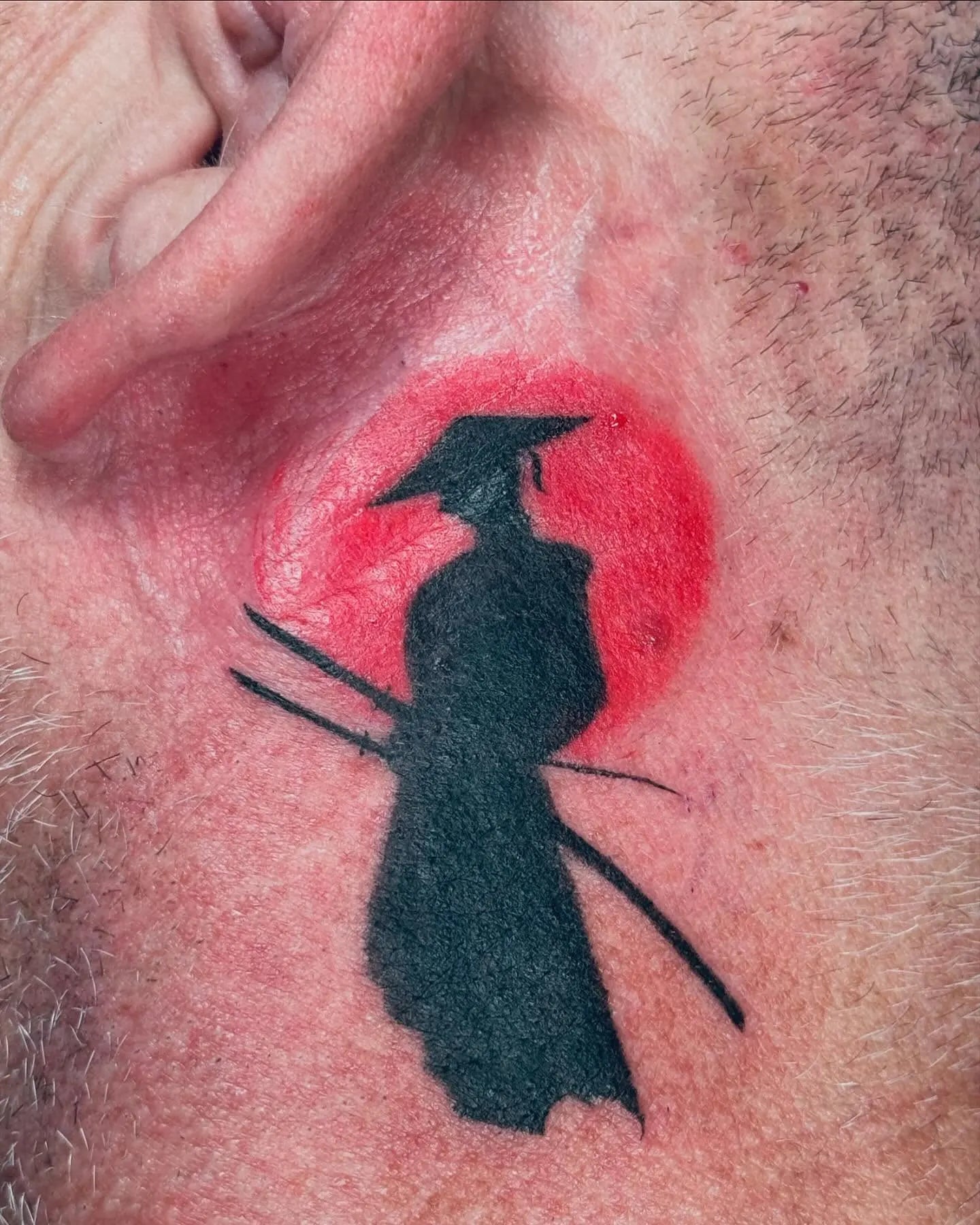 Tattoo of a black samurai silhouette figure against a red circle japanese sun behind the ear