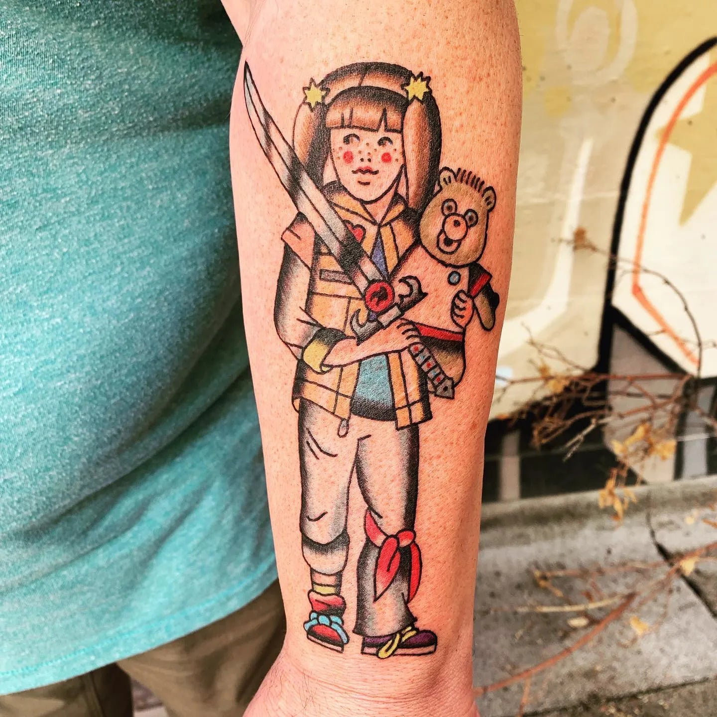 Tattoo of a girl holding a teddy bear and a sword on forearm