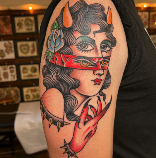 Tattoo of a stylized face with horns and a red mask on a person's arm