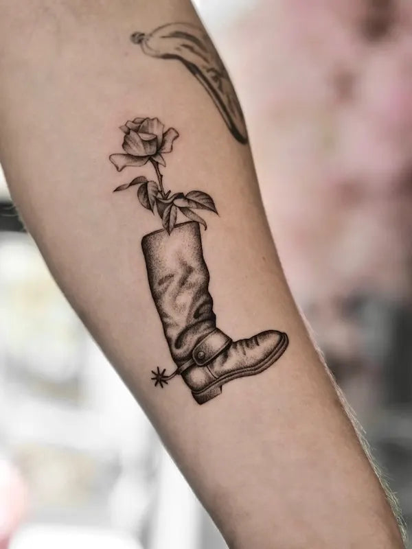 black and grey realism cowboy boot with flower tattoo on forearm